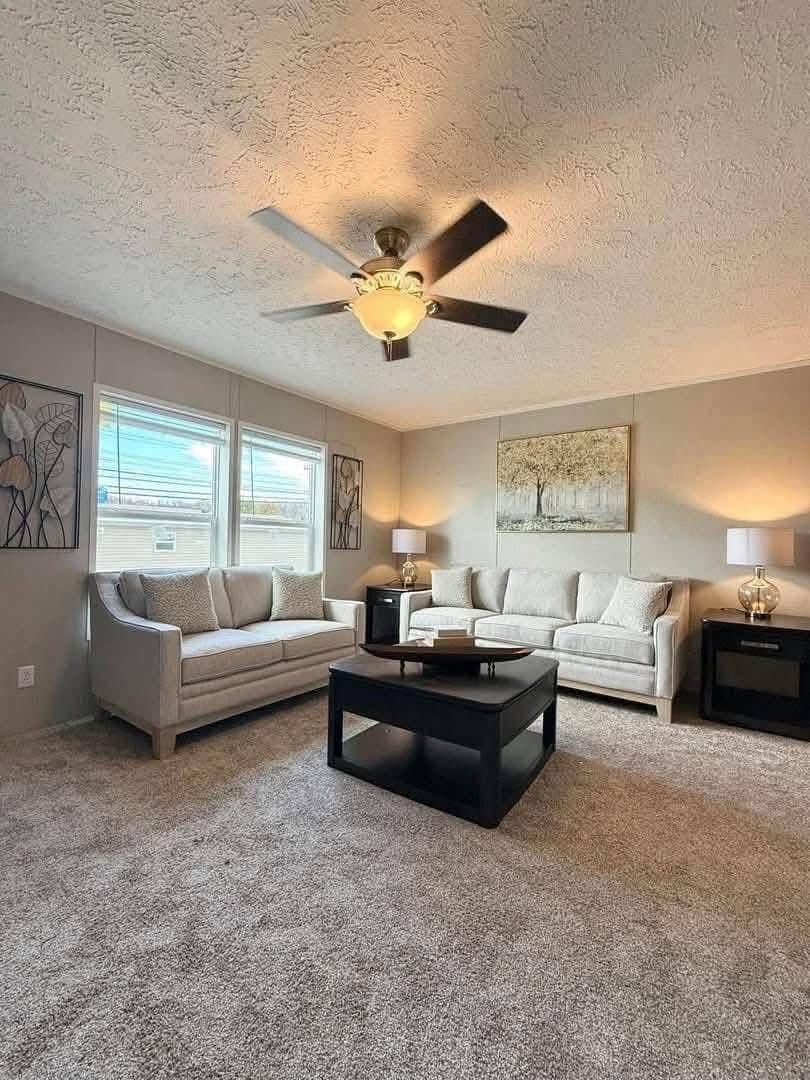 A cozy living room with two gray sofas, a black coffee table, and lamps on side tables. Art decorates the beige walls. A ceiling fan is above.