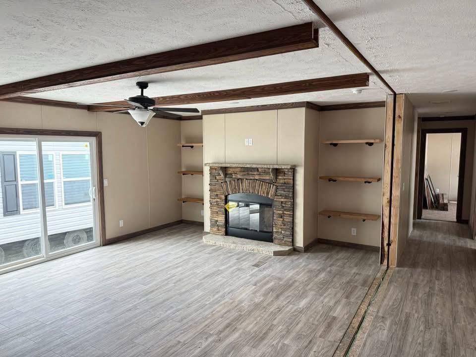 Living room with gray wood floors, stone fireplace, and wooden ceiling beams. Four built-in shelves and a ceiling fan add cozy, rustic charm.