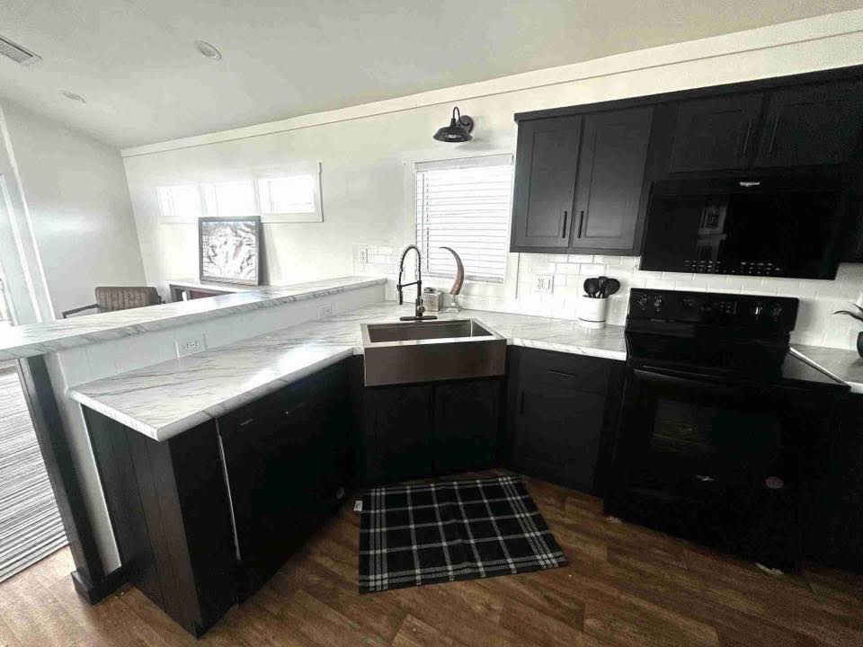 Modern kitchen with black cabinets, white marble countertops, and a farmhouse sink. Black appliances complement wood flooring and a plaid rug below.