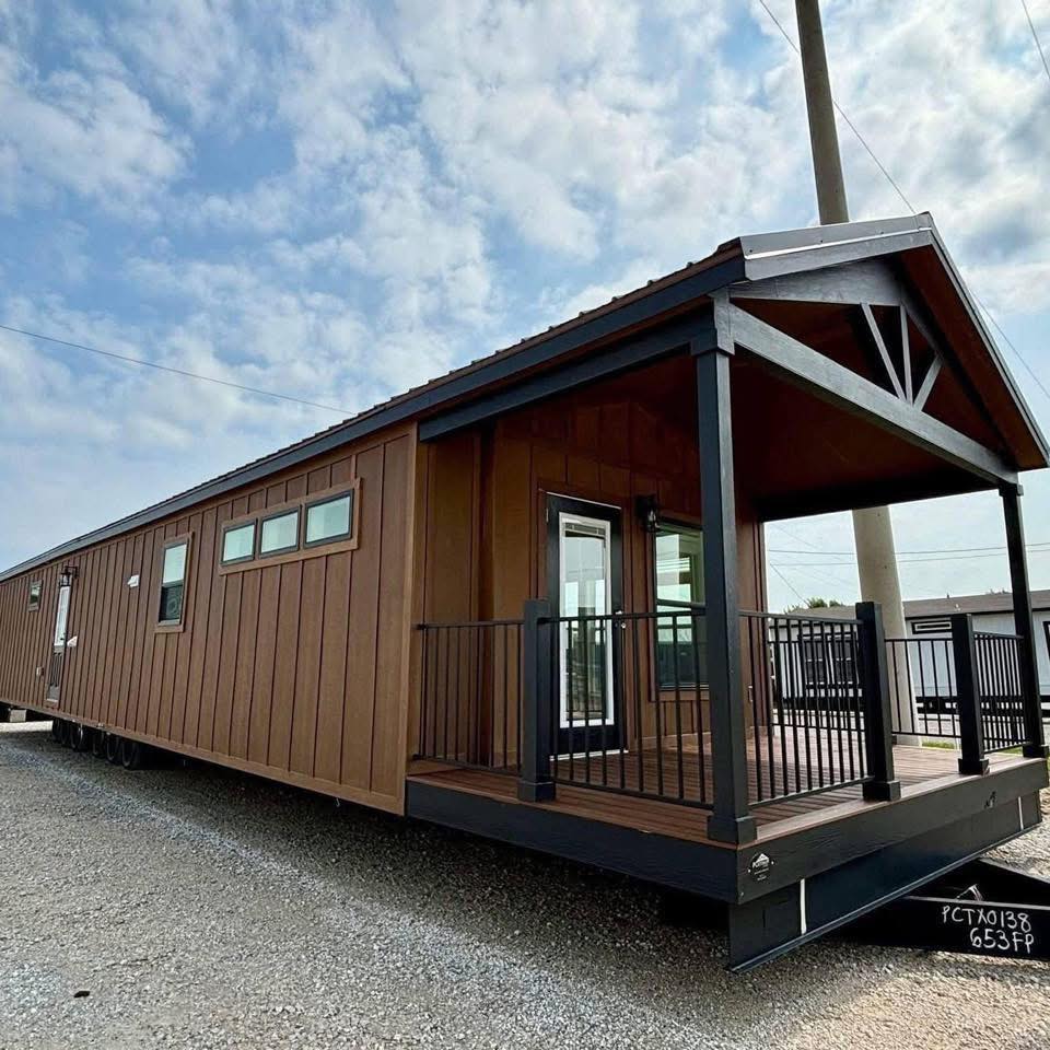 A modern brown tiny home with a gabled roof and raised porch featuring black railings. The sky is partly cloudy, adding an airy, inviting feel.