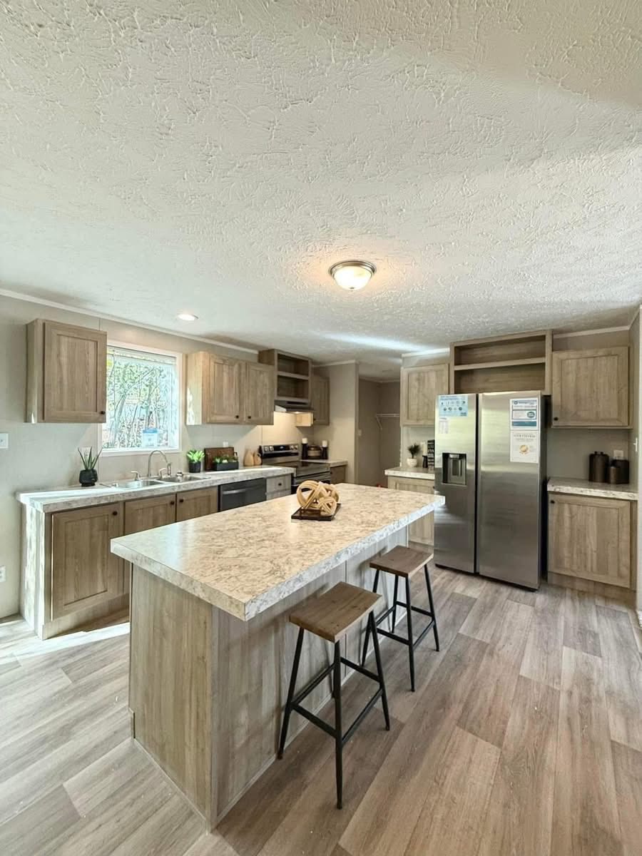 Bright, modern kitchen with wood cabinets and light countertops. An island with two stools stands central. A large window adds natural light.