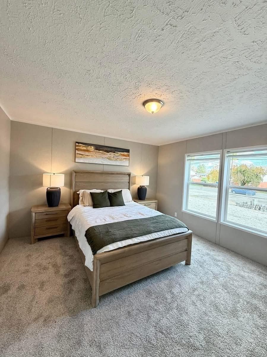 A cozy bedroom with a wooden bed, white and green bedding, and two nightstands with lamps. Large windows on the right bring in natural light.