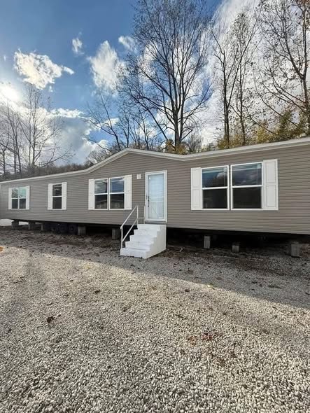 A beige mobile home with white trim sits on a gravel lot, surrounded by leafless trees under a partly cloudy sky, conveying a serene, autumnal feel.