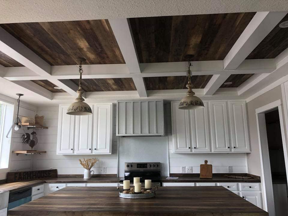 Rustic farmhouse kitchen with a wooden coffered ceiling, white cabinets, and a dark wood countertop. Two pendant lights hang over a centerpiece with candles.