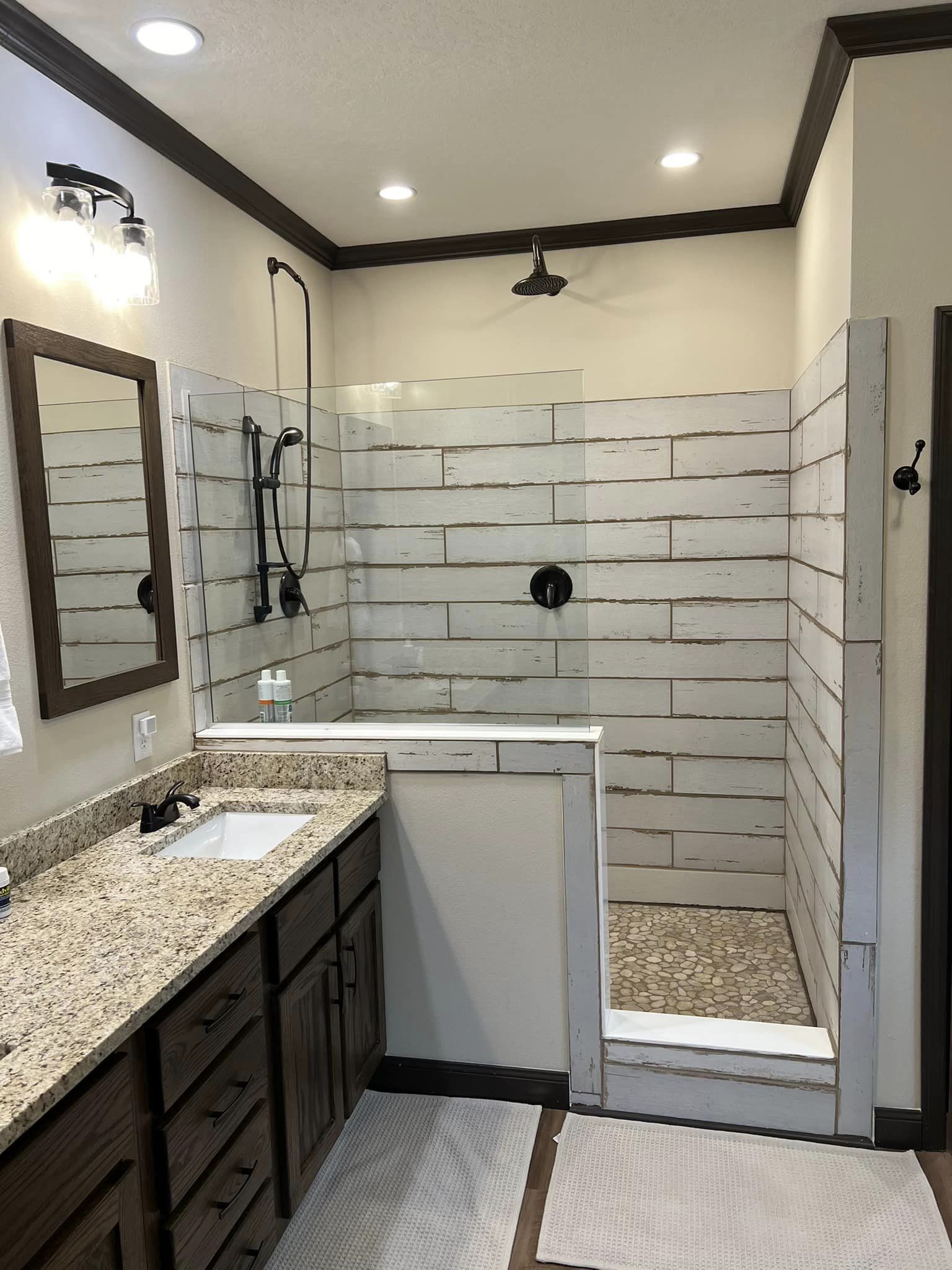 A cozy bathroom features a granite countertop with wooden cabinets, a modern shower with rustic white tile walls, a rainfall showerhead, and soft lighting.