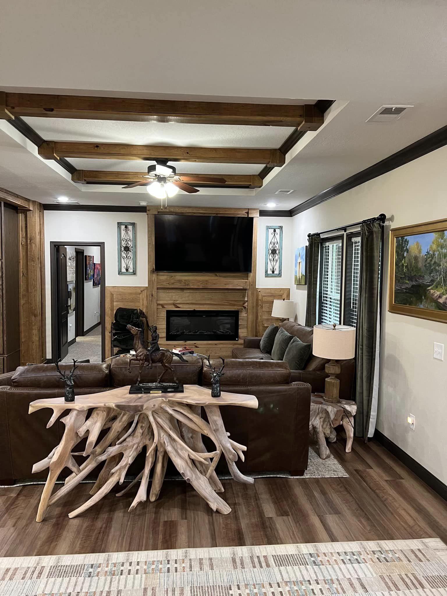 Rustic living room with wooden beams and a ceiling fan. A large TV is mounted above a fireplace. A driftwood table is behind a brown leather sofa.
