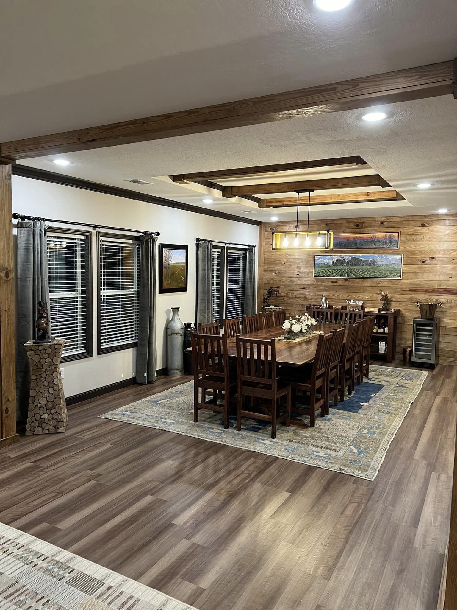 Stylish, rustic dining room with wooden floors and beams, featuring a long table for twelve, modern lighting, large landscape art, and floral centerpiece.