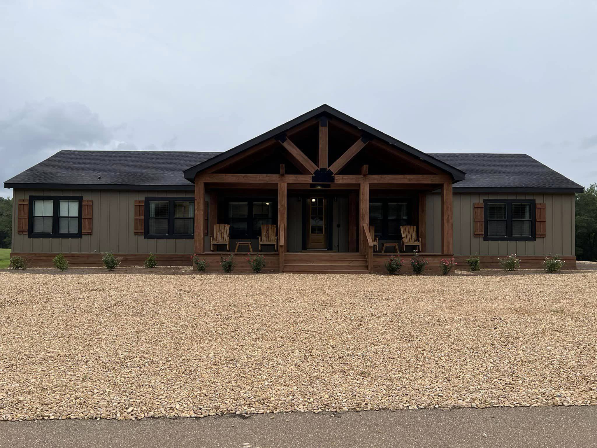Ranch-style home with a dark roof and wooden accents. The front porch has rocking chairs and a gravel pathway, creating a cozy and inviting feel.