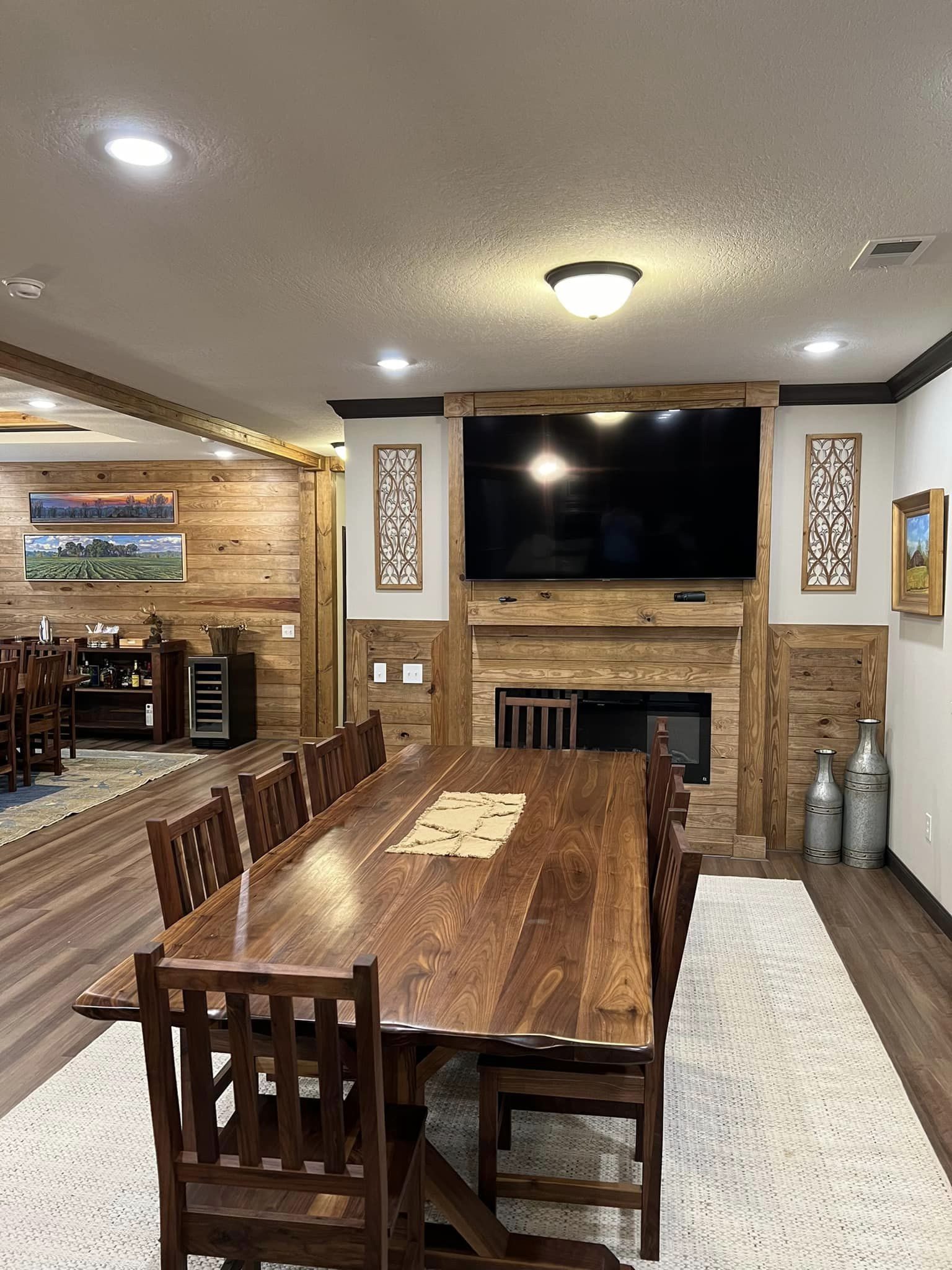 Cozy dining room with a large wooden table and chairs on a light rug. A fireplace is beneath a mounted TV with decorative panels. Warm lighting and framed art create a rustic, inviting atmosphere.