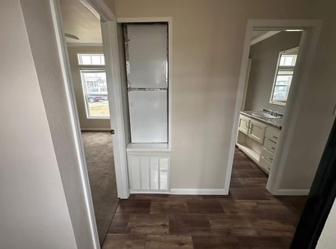 Narrow hallway with wooden flooring, leading to carpeted room on left and a bathroom with sink on right. Natural light filters through large windows.