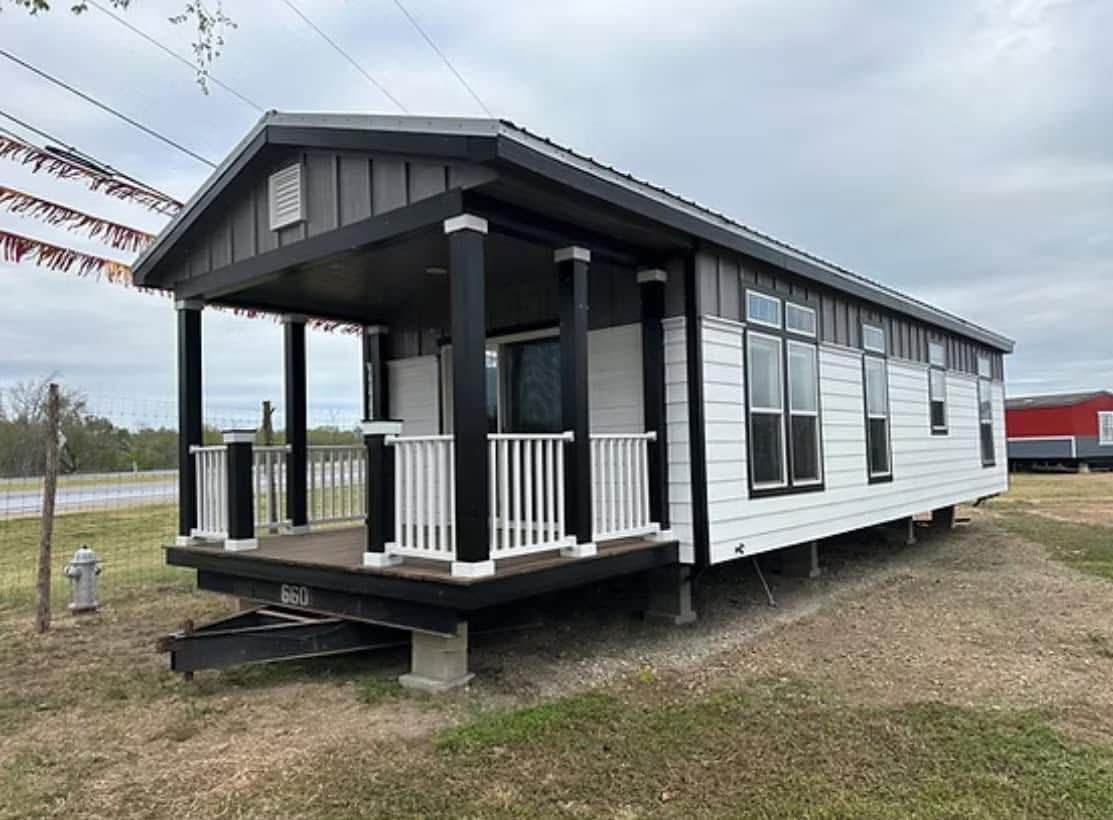 Small, elevated modular home with black pillars, a wooden porch, and white siding. It sits on a grassy lot with a cloudy sky in the background.