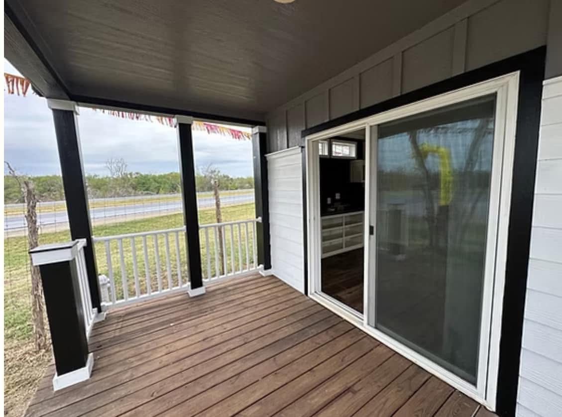 Covered porch with wooden flooring and white railings, featuring a glass sliding door leading to the interior. Overcast sky and grassy landscape in the background.