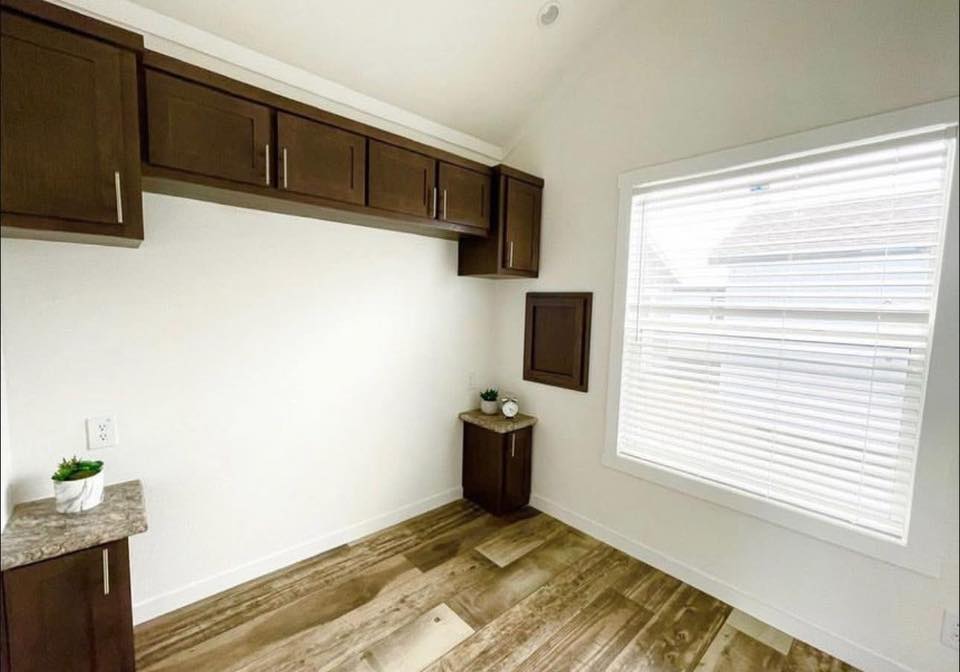 A bright, minimalist room with light wooden floors and white walls. Dark wooden cabinets line the top wall. A large window with blinds lets in natural light.