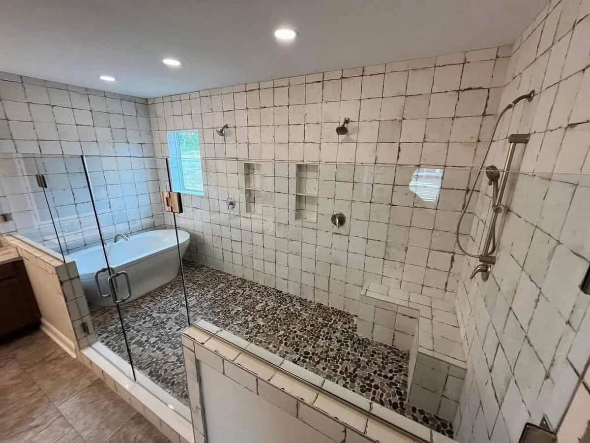 Spacious bathroom featuring white tiled walls, a glass-enclosed shower with pebble flooring, multiple showerheads, and a standalone bathtub. Bright and modern.