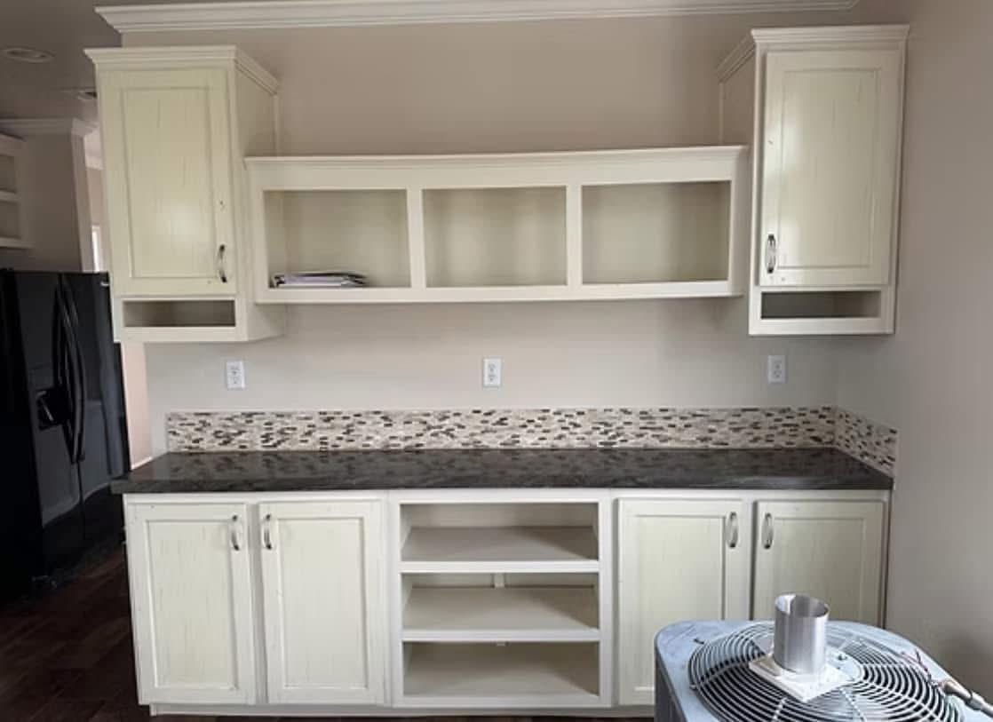 Cream-colored kitchen cabinets with open shelving and a dark countertop. Mosaic tile backsplash adds texture. Calm, neutral tones.
