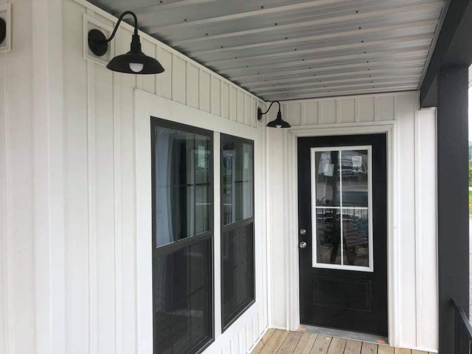 Covered porch with a black door, large window, and two black sconces on white siding. The wooden floor adds warmth to the modern design.