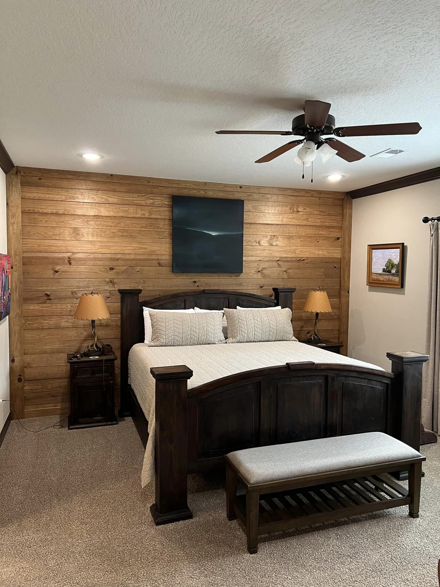 Cozy bedroom with a wooden accent wall, dark wood bed frame, and beige bedding. Two side tables with lamps, a bench, and a ceiling fan add warmth.