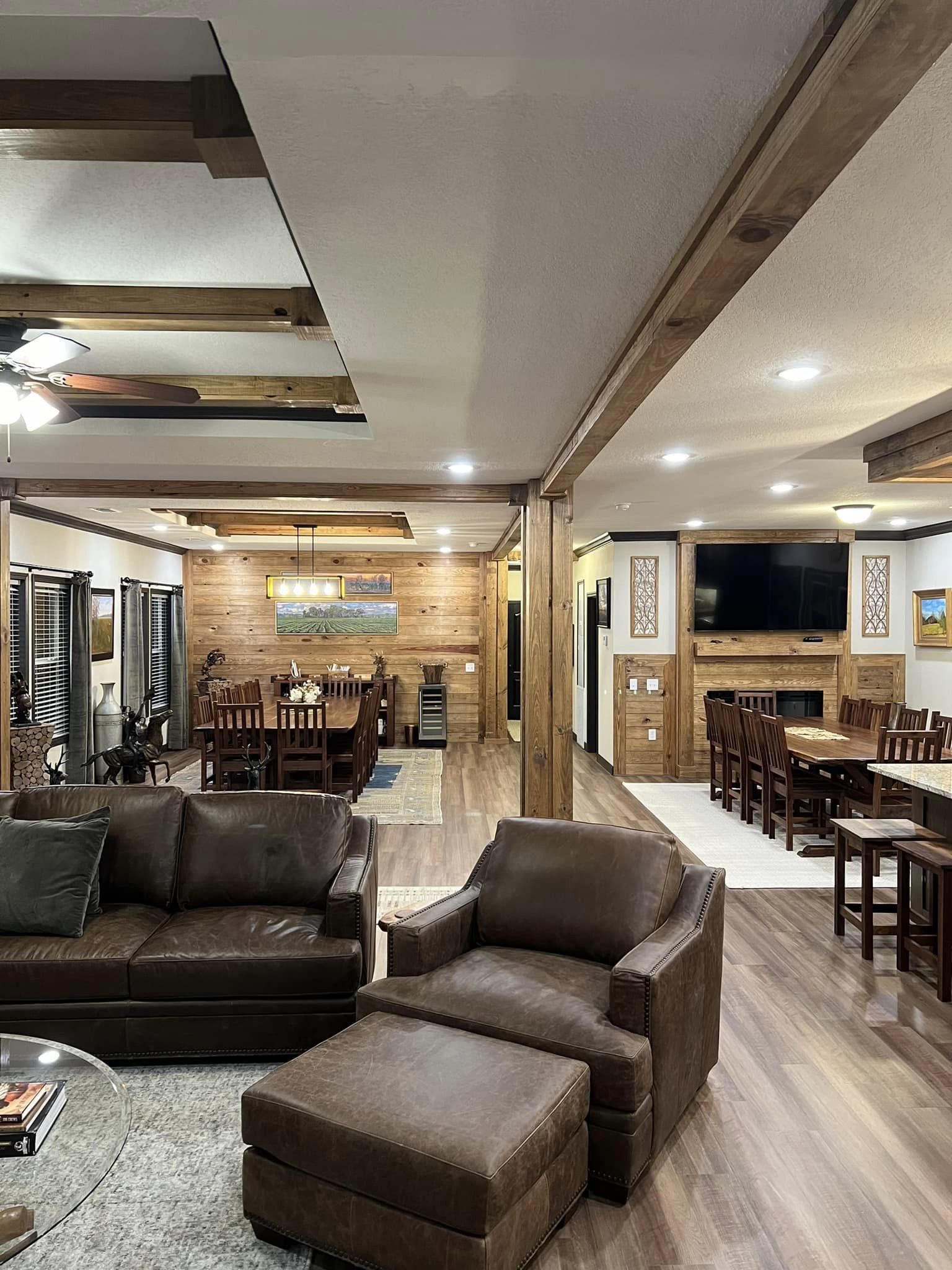 Spacious living room with leather sofa and armchair, leading to a wooden dining area. Warm lighting, rustic decor, and wooden beams create a cozy ambiance.