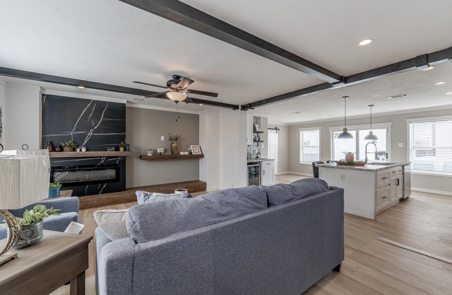 Modern open-concept living room with gray sofa, sleek kitchen island, wood accents, large windows, and black beam ceiling. Cozy and inviting.