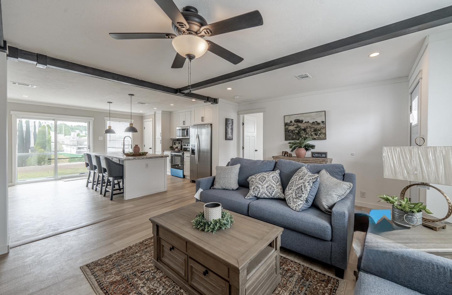 Modern living area with blue sofa, patterned cushions, and wooden coffee table. Open kitchen with island, ceiling fan, and natural light from large windows. Cozy and inviting atmosphere.
