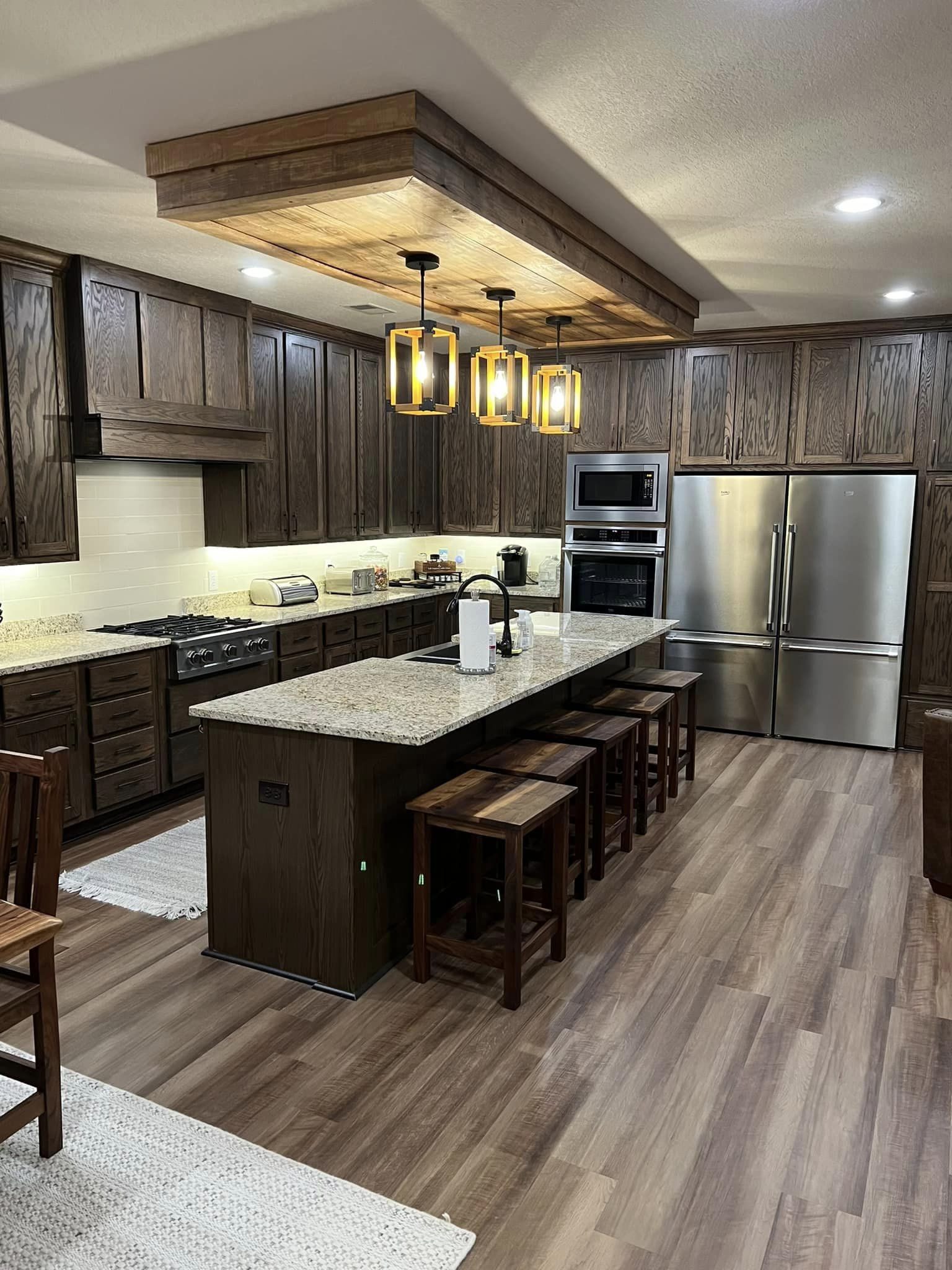 Spacious kitchen with wooden cabinets, granite island with four stools, stainless steel appliances, and three pendant lights, creating a cozy ambiance.
