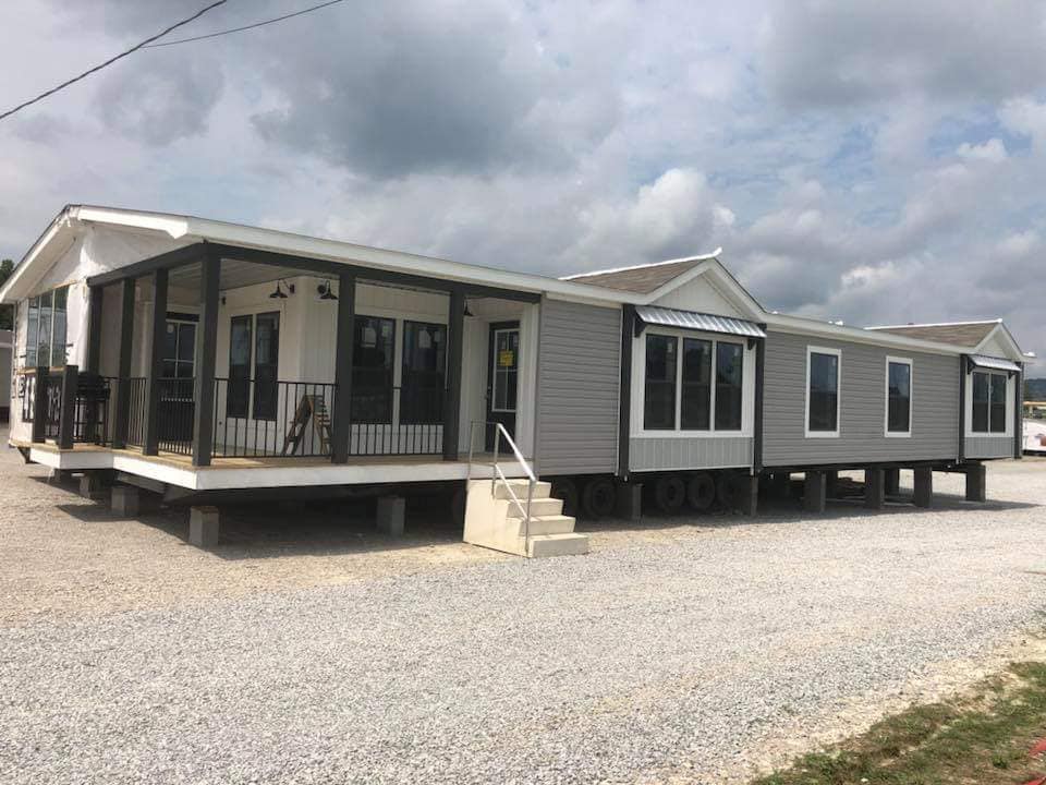 Large modular home with a porch, sitting on gravel under a cloudy sky. The structure is elevated with stairs, conveying a modern and spacious feel.