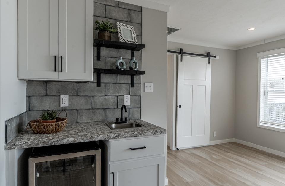 A modern kitchenette with a gray tile backsplash, granite countertop, and small sink. Open shelves hold decor. A barn door and window are in view.