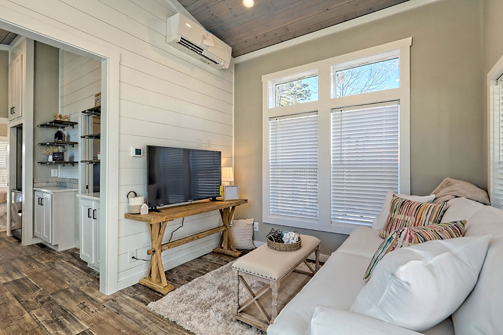 Cozy living room with natural light. Features white sofa with colorful pillows, wooden TV stand, and decorative items. Warm, inviting ambiance.