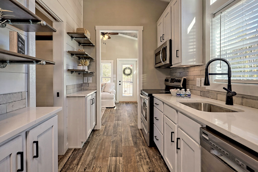 Narrow kitchen with white cabinets, stainless steel appliances, and dark wood flooring. Sunlight streams through a window, creating a cozy ambiance.