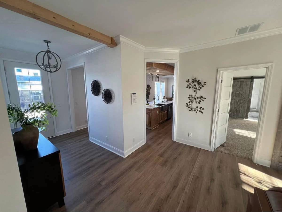 Bright, modern living space with wooden floors and white walls. Features include a leafy wall decoration, pendant light, and open doors leading to a kitchen and sunlit room. Calm, inviting atmosphere.