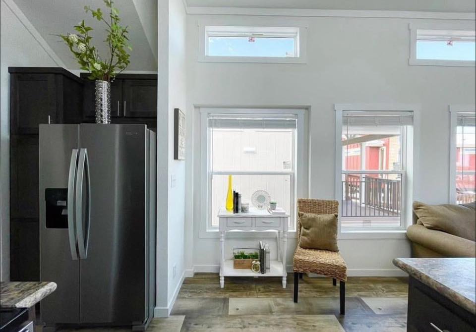 A modern kitchen corner with a stainless steel fridge and a wicker chair beside a white table. Large windows let in natural light, creating a cozy ambiance.