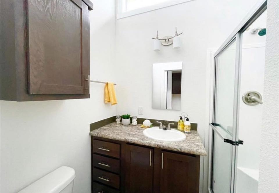 Compact, bright bathroom with dark wood cabinets, a granite countertop with a sink, a mirror, yellow towel, shower with glass door, and potted plants.