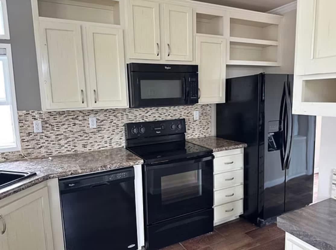A modern kitchen with beige cabinets, a black fridge, stove, microwave, and dishwasher. Mosaic backsplash and brown countertops add a warm touch.