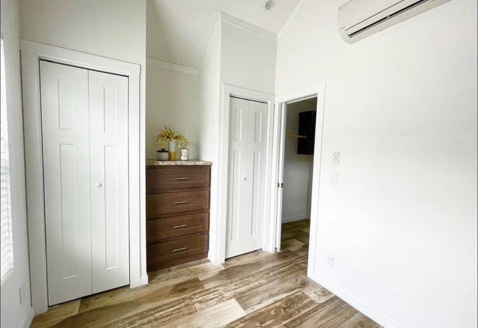 A small, minimalist room with white walls, wooden flooring, and two white doors. A dark wooden dresser with plants and a bottle adds warmth.