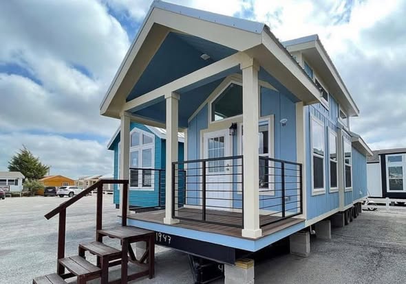 A charming blue tiny house with a white-trimmed porch and stairs. It sits in a sunny lot under a partly cloudy sky, conveying a cozy and welcoming feel.