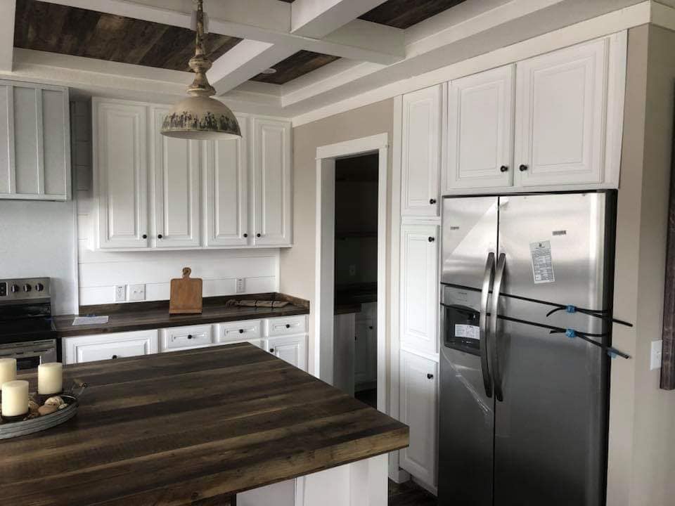 Bright kitchen with white cabinets, stainless steel fridge, and wooden countertops. Rustic island holds candles; pendant light hangs above. Cozy and inviting.