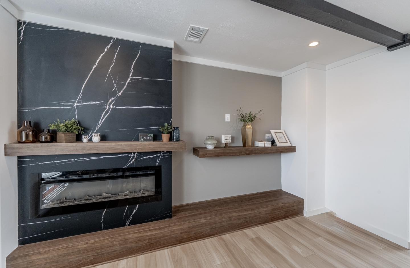 Modern living room with a black marble fireplace, wooden mantel topped with plants and decor. Neutral walls, floating shelf with additional plants and items.