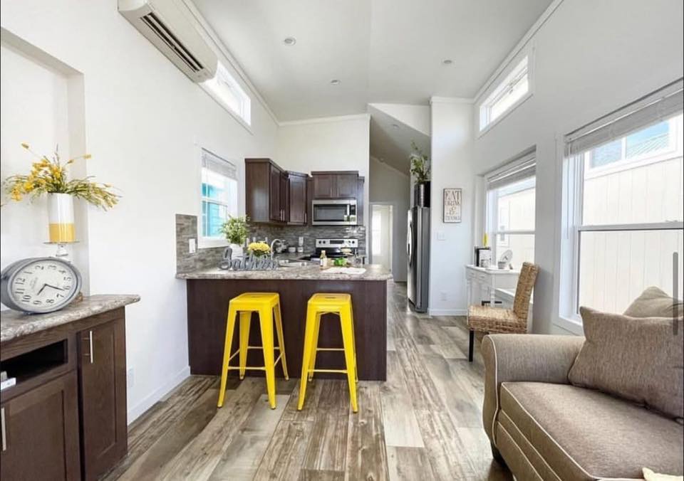 Modern kitchen and living area with white walls and wood flooring. Features include dark cabinets, bright yellow bar stools, and a beige sofa.