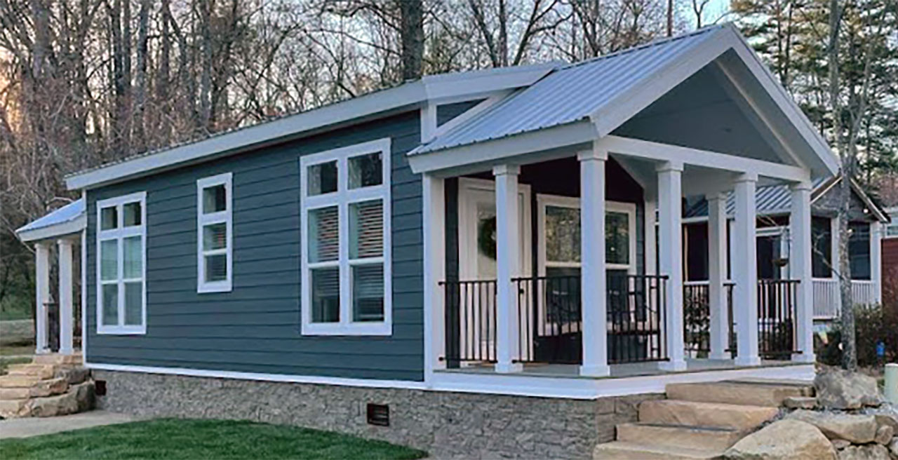 A modern tiny house with blue siding, large windows, and a covered porch. Set on a stone foundation, it is surrounded by a wooded area and lawn.