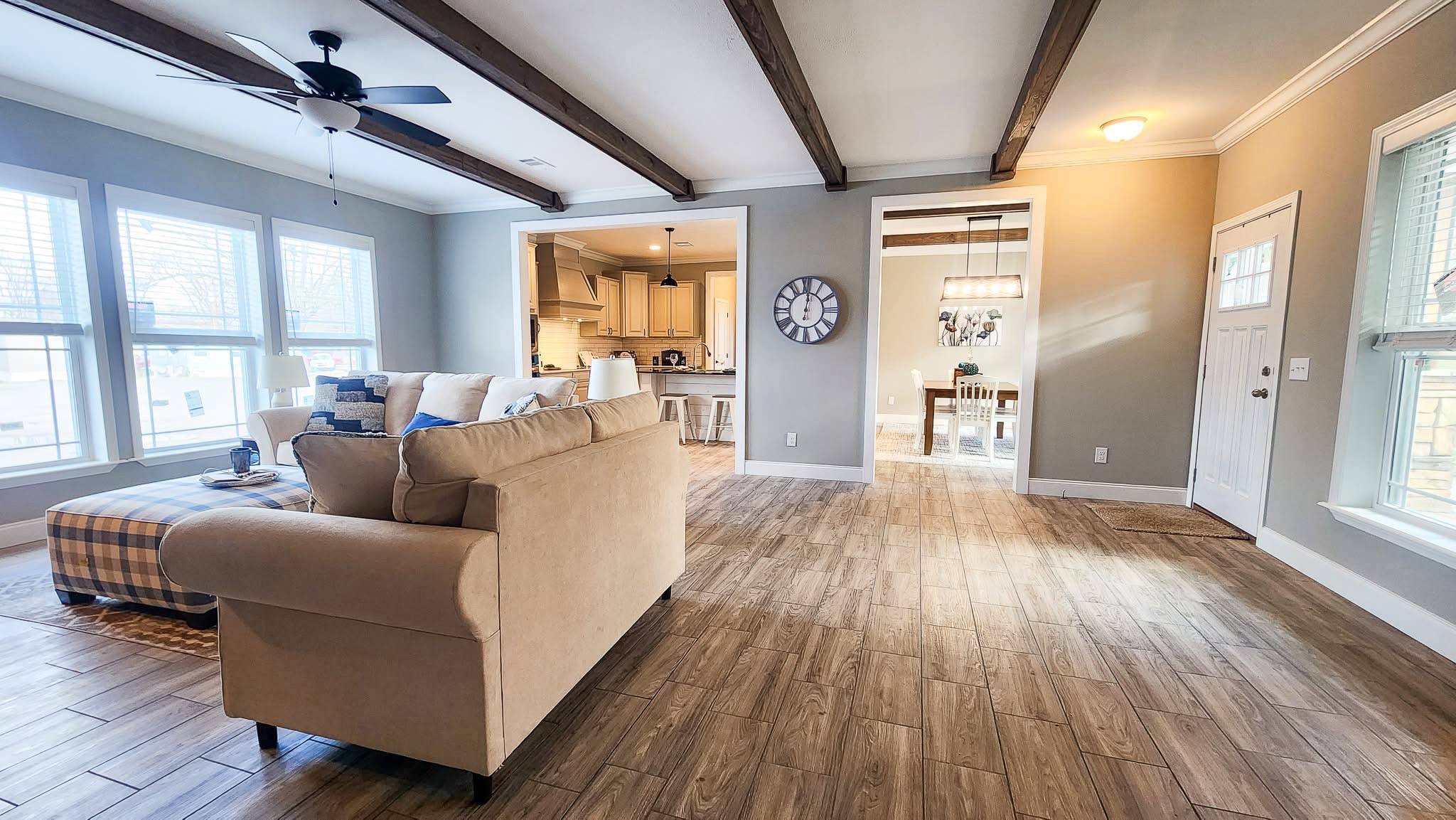 Spacious living room with wood beams and light wood flooring, featuring a beige sofa. Large windows provide natural light. Kitchen and dining areas visible.