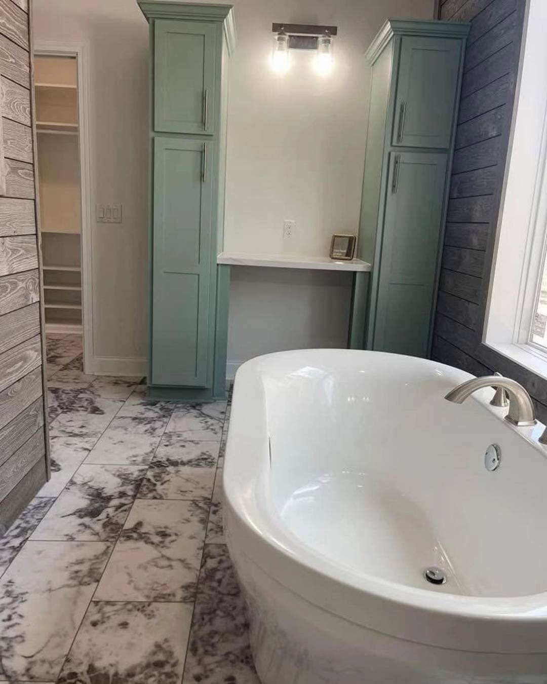 Luxurious bathroom with a white freestanding tub, marble-patterned floor tiles, mint green cabinets, wood accents, and a soft ambient light fixture.