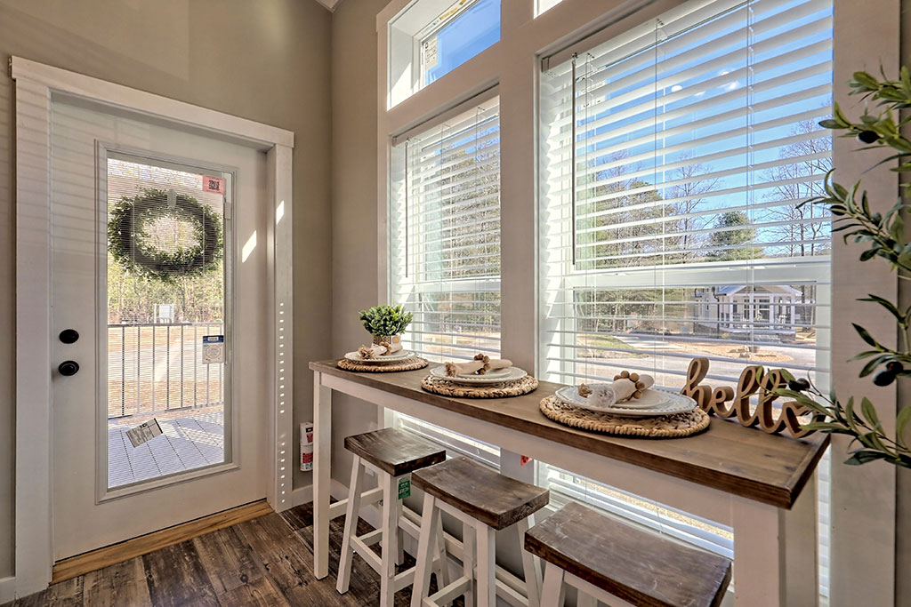 Cozy breakfast nook with tall windows, a wooden table set with round placemats, white stools, and a decorative "hello" sign, bathing in warm sunlight.