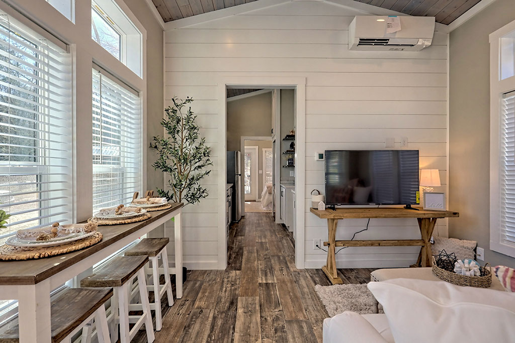 Bright, cozy living space with white walls and wood floors. A narrow bar with stools is by the window, a TV on a rustic stand, and a leafy plant adds greenery.