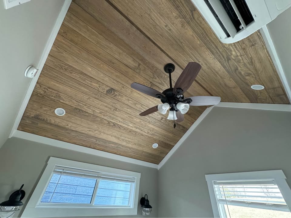 Wood-paneled ceiling with a central ceiling fan and lights. Two windows, wall lamps, and an air conditioner create a cozy, modern room ambiance.