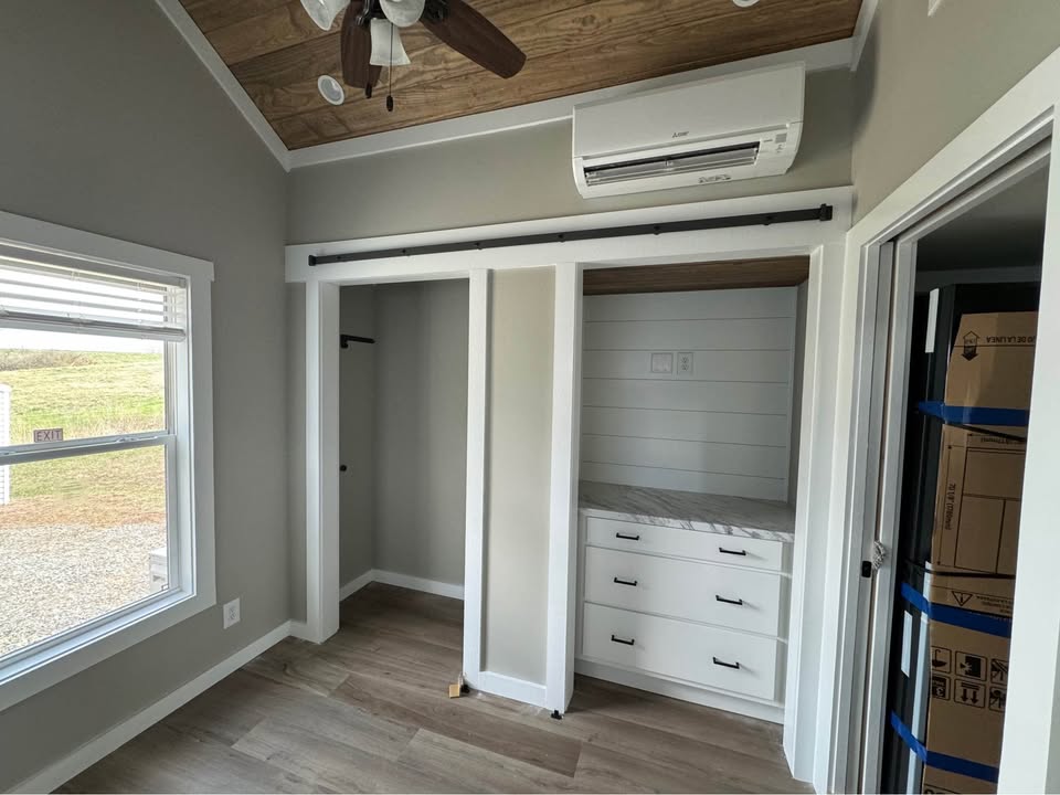 Cozy room with wooden ceiling, fan, and a window. It features an open closet with drawers and hanger space, and a wall-mounted air conditioner.
