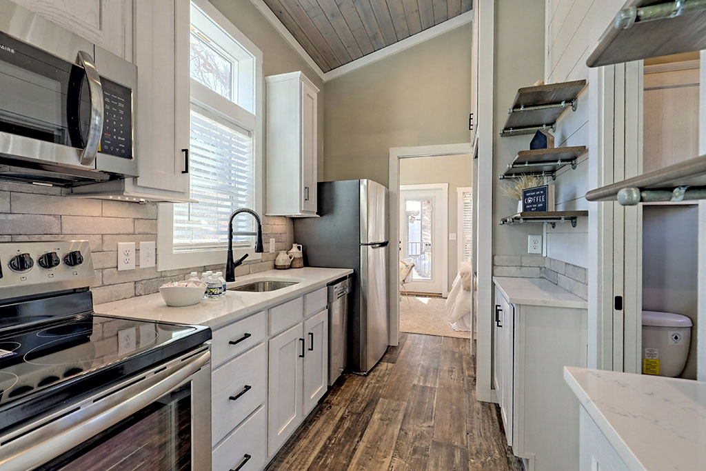 A modern kitchen with white cabinets, stainless steel appliances, a black faucet, and wood flooring. Shelves hold decor items, creating a cozy ambiance.