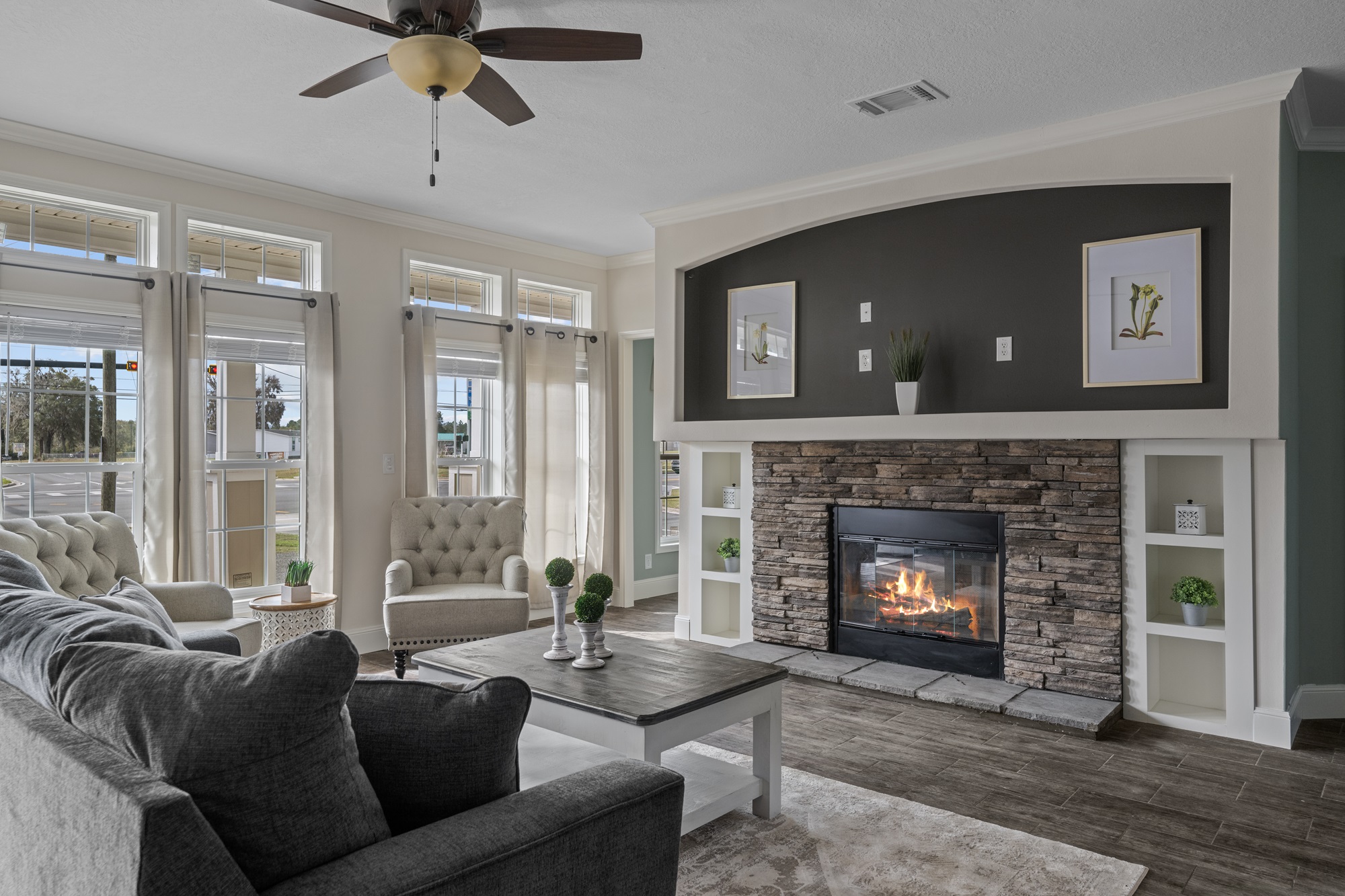 Modern living room with stone fireplace, gray couches, and large windows. Natural light fills the space, creating a cozy and inviting atmosphere.