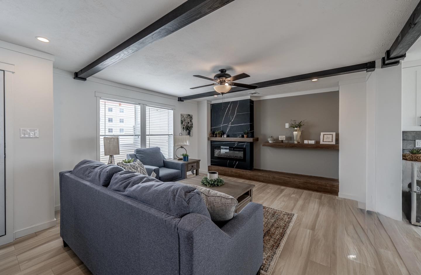 Modern living room with a cozy vibe; features a blue sofa, large windows, ceiling fan, and a fireplace with a dark accent wall and wooden mantle.