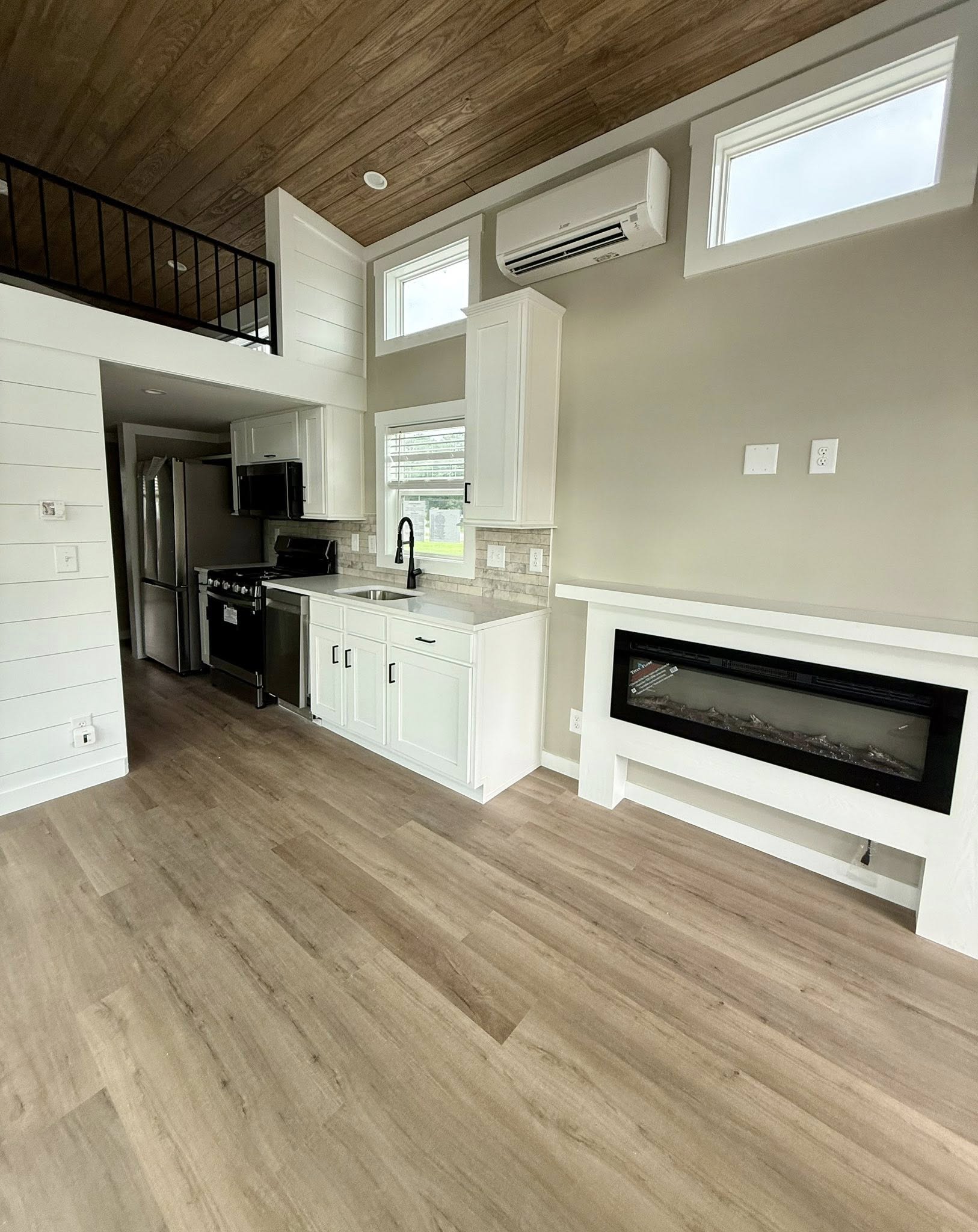 Modern tiny home interior with wood flooring, white cabinets, and stainless steel kitchen appliances. Features a wall-mounted fireplace and a loft.