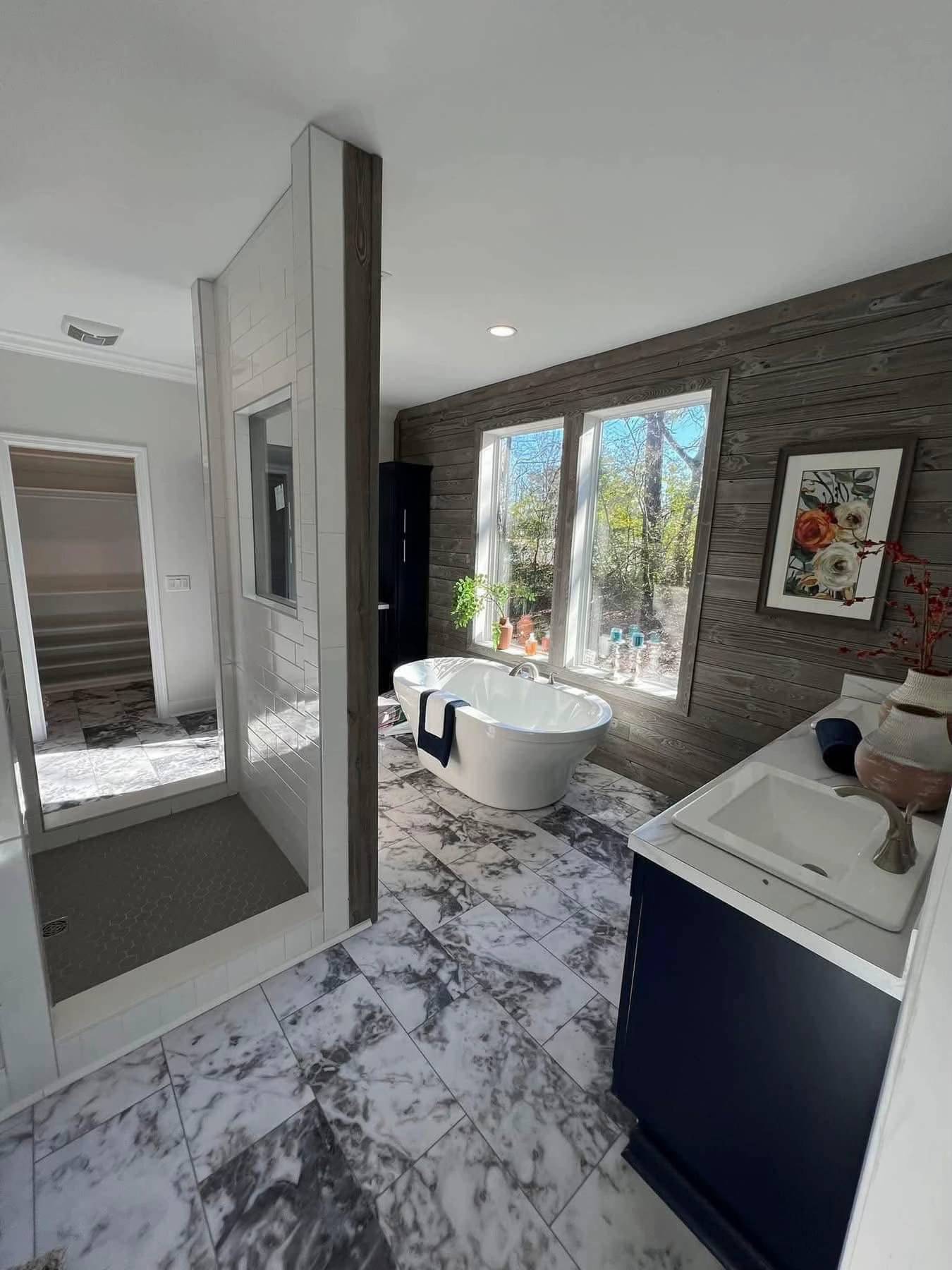 Luxurious bathroom with marble tiles, a standalone white tub by large windows, a sleek glass shower, and a navy vanity. Bright, airy ambiance.